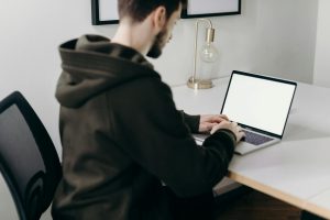 A young man in a hoodie working on a laptop at a minimalist home office desk.