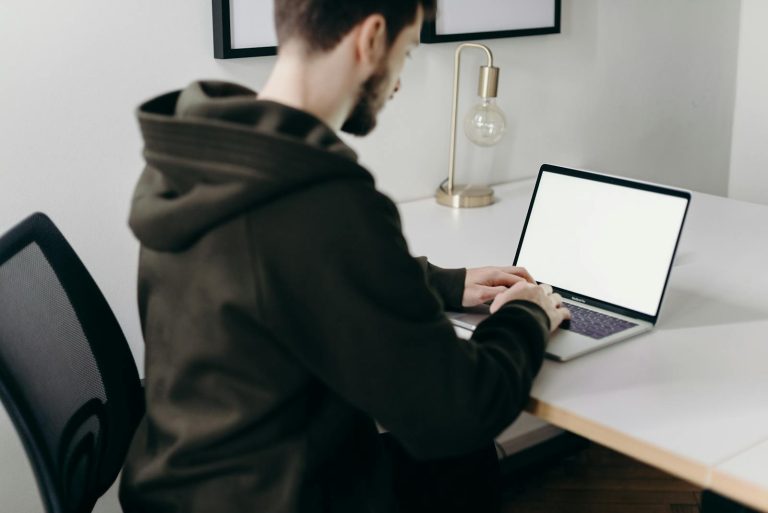 A young man in a hoodie working on a laptop at a minimalist home office desk.